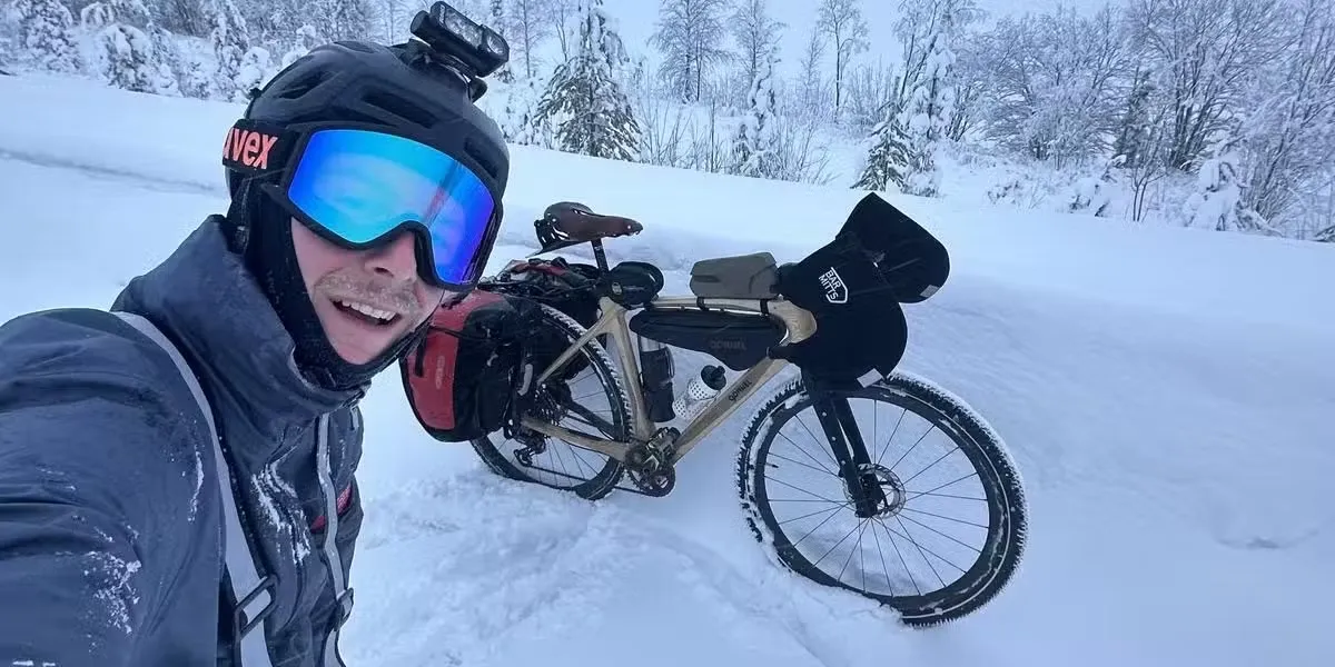 Baptiste Ecalle en selfie dans la neige, lunettes de ski sur le nez, à côté de son vélo en bois chargé de sacoches