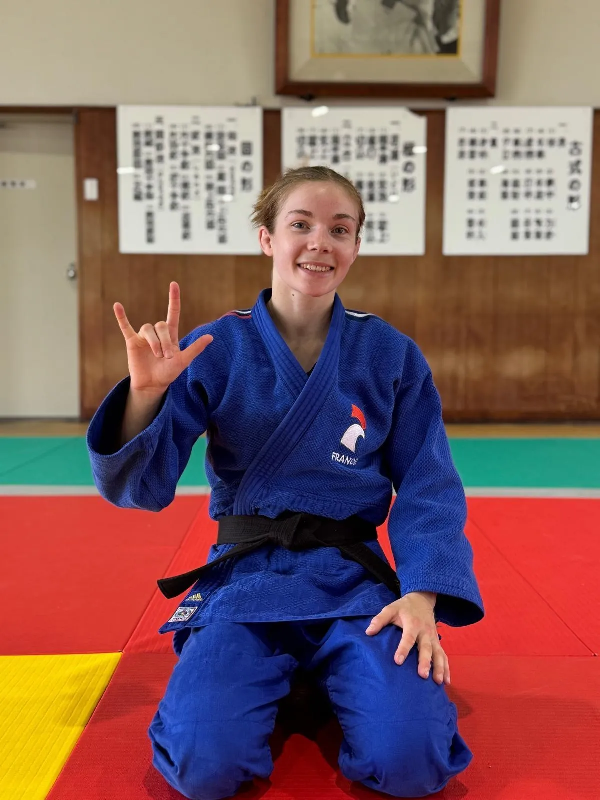 Clara Mermet, en kimono bleu de l'équipe de France, sourit à genoux sur le tatami en faisant le signe de la victoire