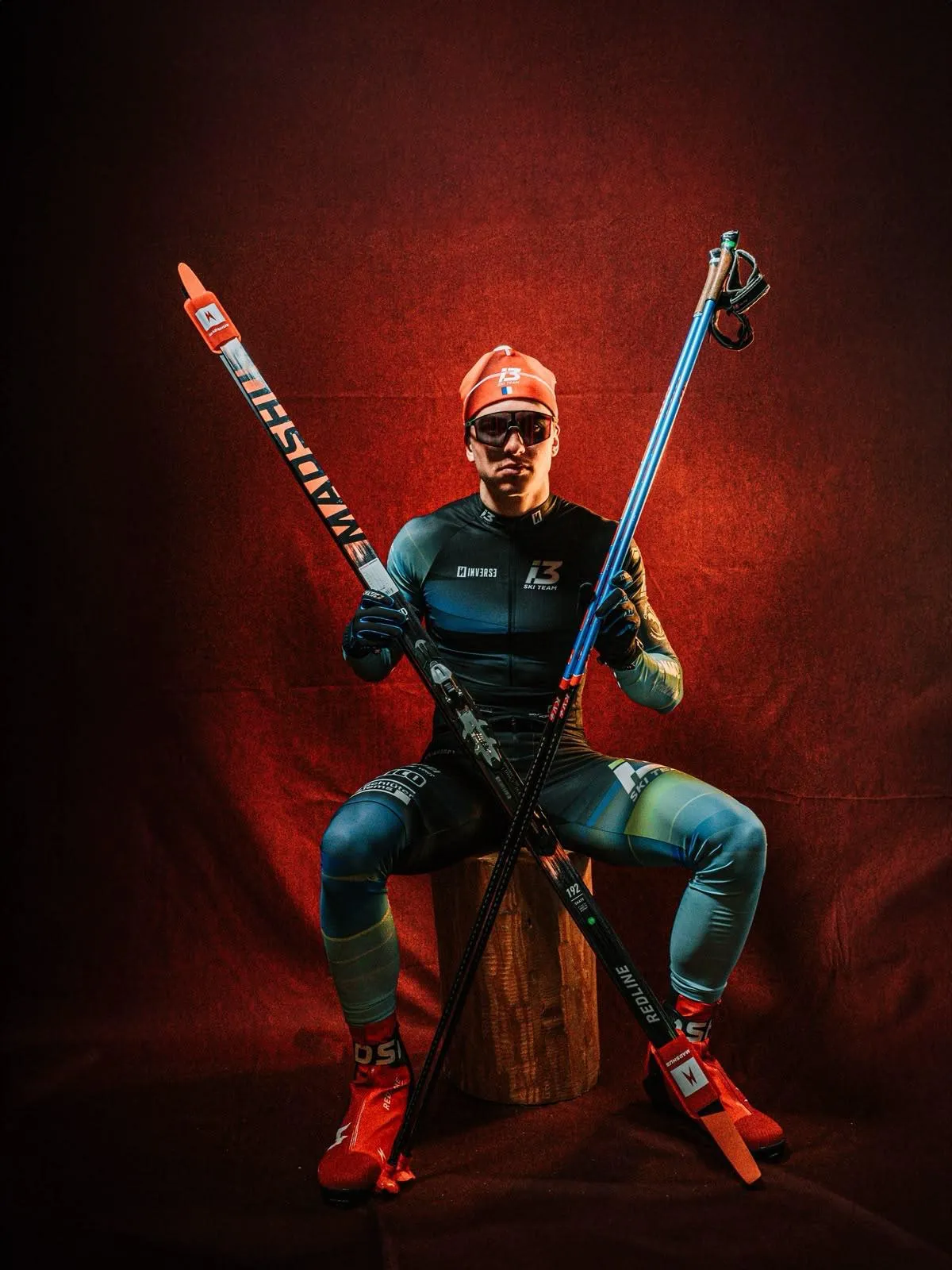 Noa Genest, bonnet orange et lunettes de soleil, assis sur une bûche, croise ses skis et ses bâtons devant un drapé rouge